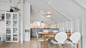A spacious dining area with a wooden table and white chairs, a white cabinet with glass doors, and a kitchen in the background.