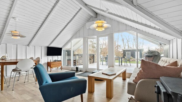 A spacious living area with a high ceiling, wooden flooring, and a fireplace. There is a television mounted on the wall, a dining table with chairs, and a sofa with cushions.