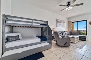 Un lit superposé avec un bureau intégré, un canapé et un ventilateur de plafond, dans une pièce bien éclairée et dotée d’une grande fenêtre.