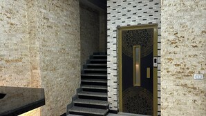 Un interior moderno con una escalera que lleva a una puerta oscura con pomo dorado, rodeada de paredes con textura y suelo de baldosas.