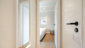 Um corredor que dá para um quarto, com uma cama, um banquinho pequeno e um quadro na parede.