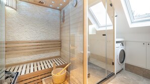 Uma sauna com banco de madeira, balde de madeira e box de chuveiro em vidro.