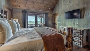 A wooden cabin bedroom with a large bed, a TV mounted on the wall, and a view of the outdoors through a sliding door.