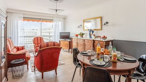 A modern living room with a dining area, featuring a large window with curtains, a television, and a dining table set with breakfast items. - Appartement Leroy, balcon - Welkeys (Clichy)