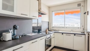 A modern kitchen with white cabinets, a black countertop, a coffee machine, and a window with a view of buildings. - Appartement Leroy, balcon - Welkeys (Clichy)