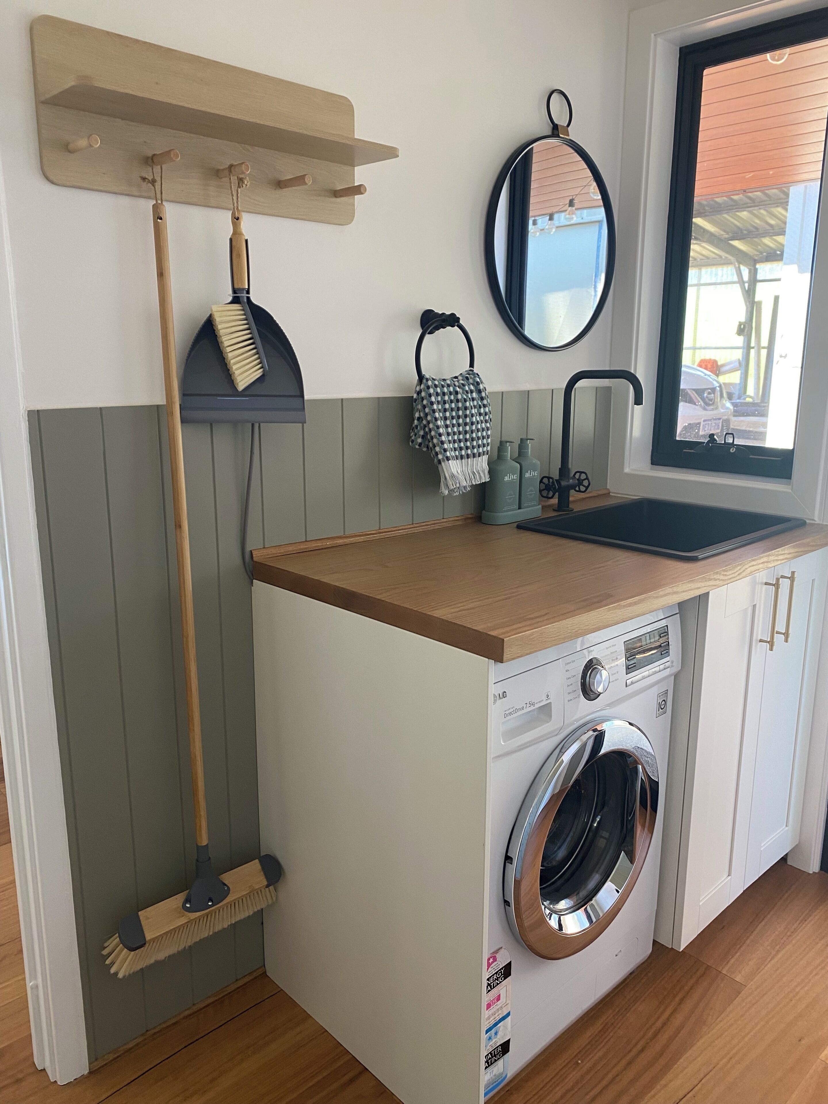 A laundry area with a washing machine, a broom, a hanging towel, and a window with a view.