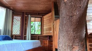 Eine Holzhütte mit einem großen Baumstamm in der Mitte, zwei Betten, einem Deckenventilator und einem farbenfrohen Streifen Teppich.