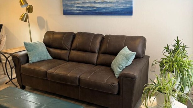 A brown leather sofa with two cushions, a wooden coffee table, a potted plant, and a painting of sailboats on the wall.