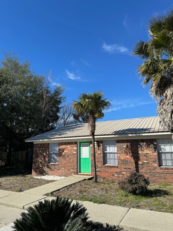 A single-story brick house with a green door, a small front yard, and a palm tree.