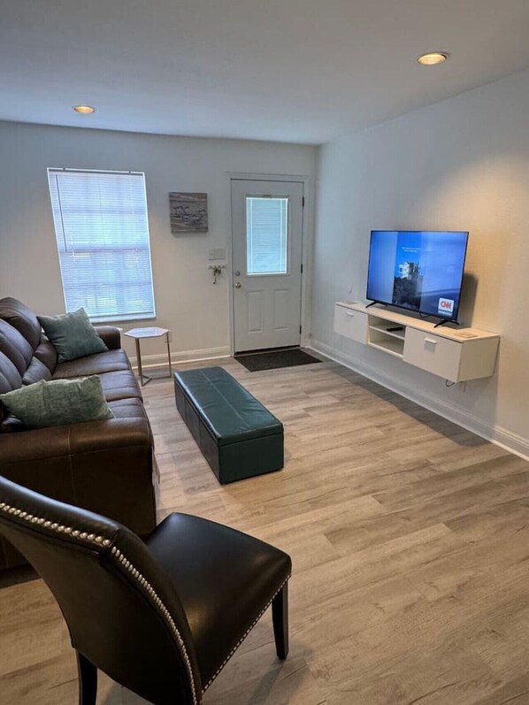 A living room with a brown leather sofa, a green ottoman, a white TV stand, and a door with a window. - Affordable Townhome in Gulf Breeze Florida is ready for guest. (Gulf Breeze)