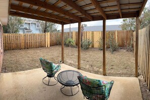 A covered patio with two metal chairs and a table, surrounded by a wooden fence and sparse vegetation. - Affordable Townhome in Gulf Breeze Florida is ready for guest. (Gulf Breeze)