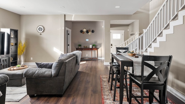 Un salon moderne avec un canapé d’angle gris, un coin repas avec une table et des chaises en bois, et un escalier menant à un autre niveau.