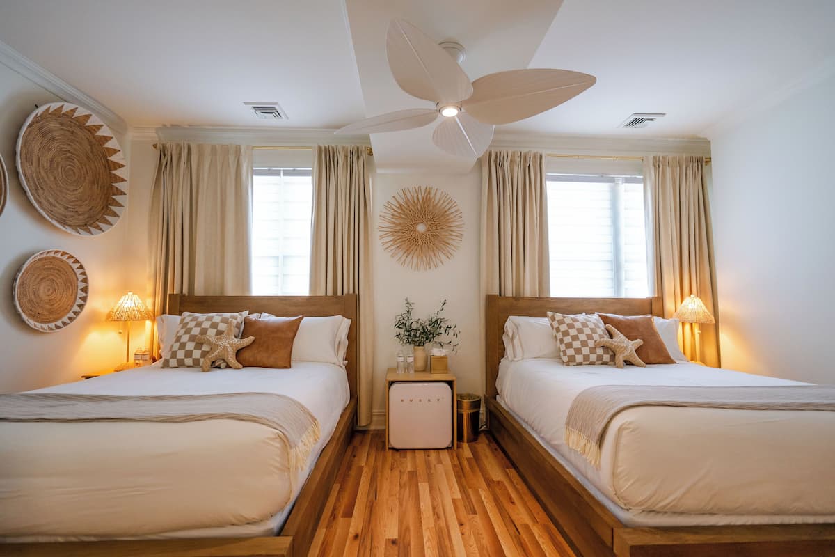 A hotel room with two beds, wooden flooring, ceiling fan, large windows with curtains, wall decor, and bedside lamps.