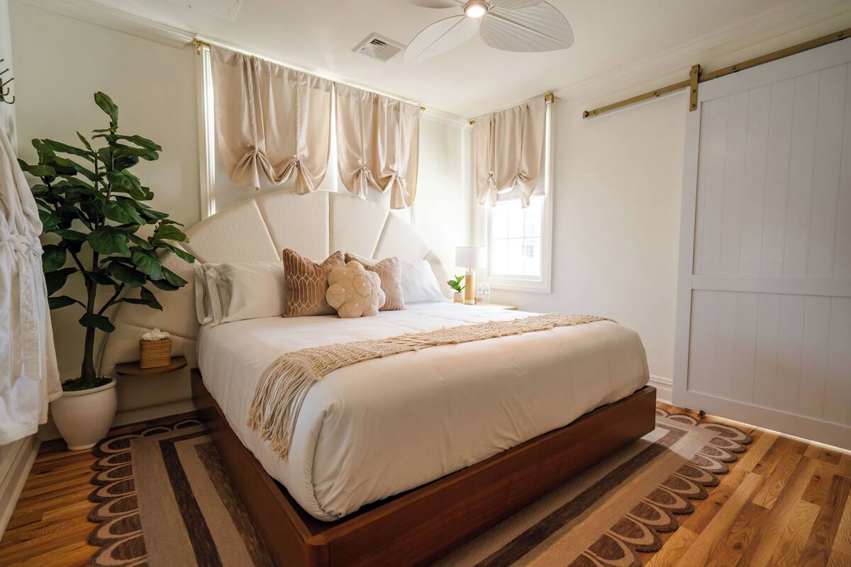 A bedroom with a wooden bed, a potted plant, a bedside table, and a large window with curtains.