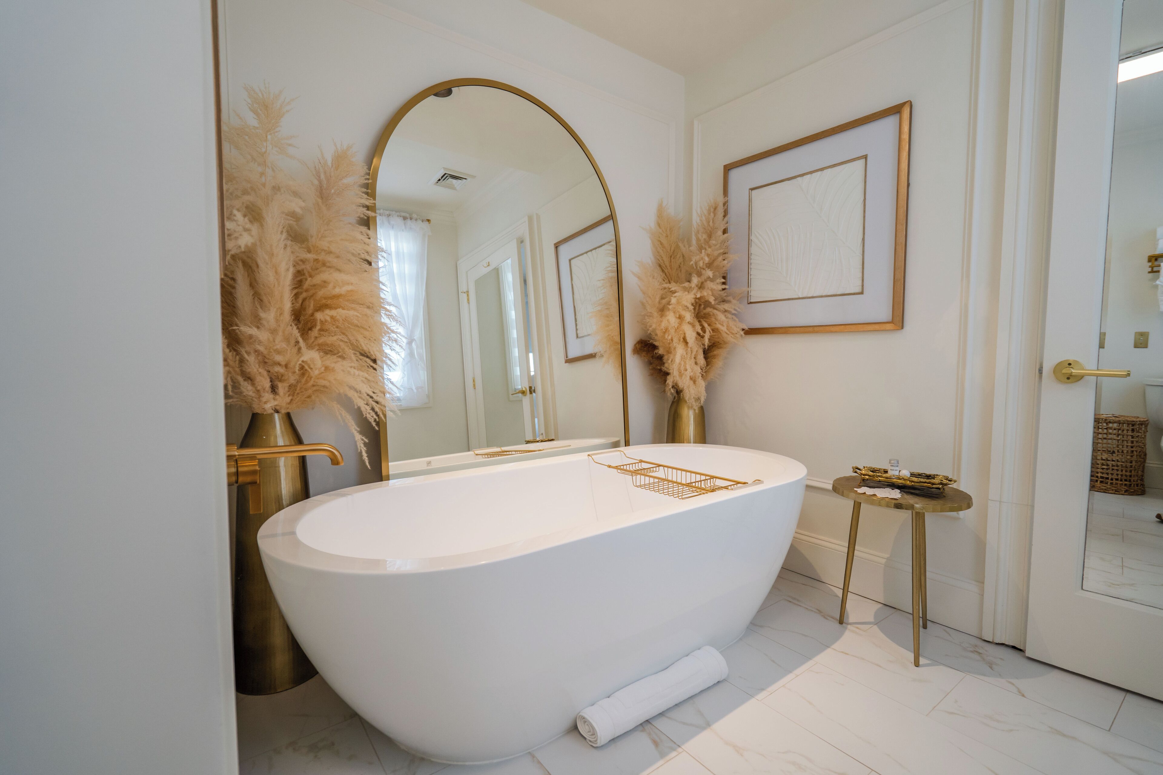 A modern bathroom with a freestanding bathtub, a large mirror, pampas grass decorations, and a small side table.
