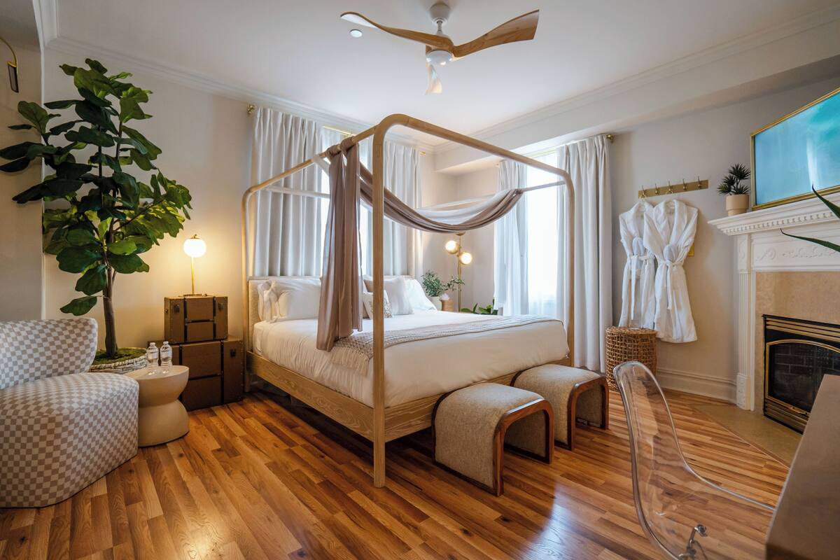 A four-poster bed with a canopy, a large plant, a fireplace, and a checkered armchair.