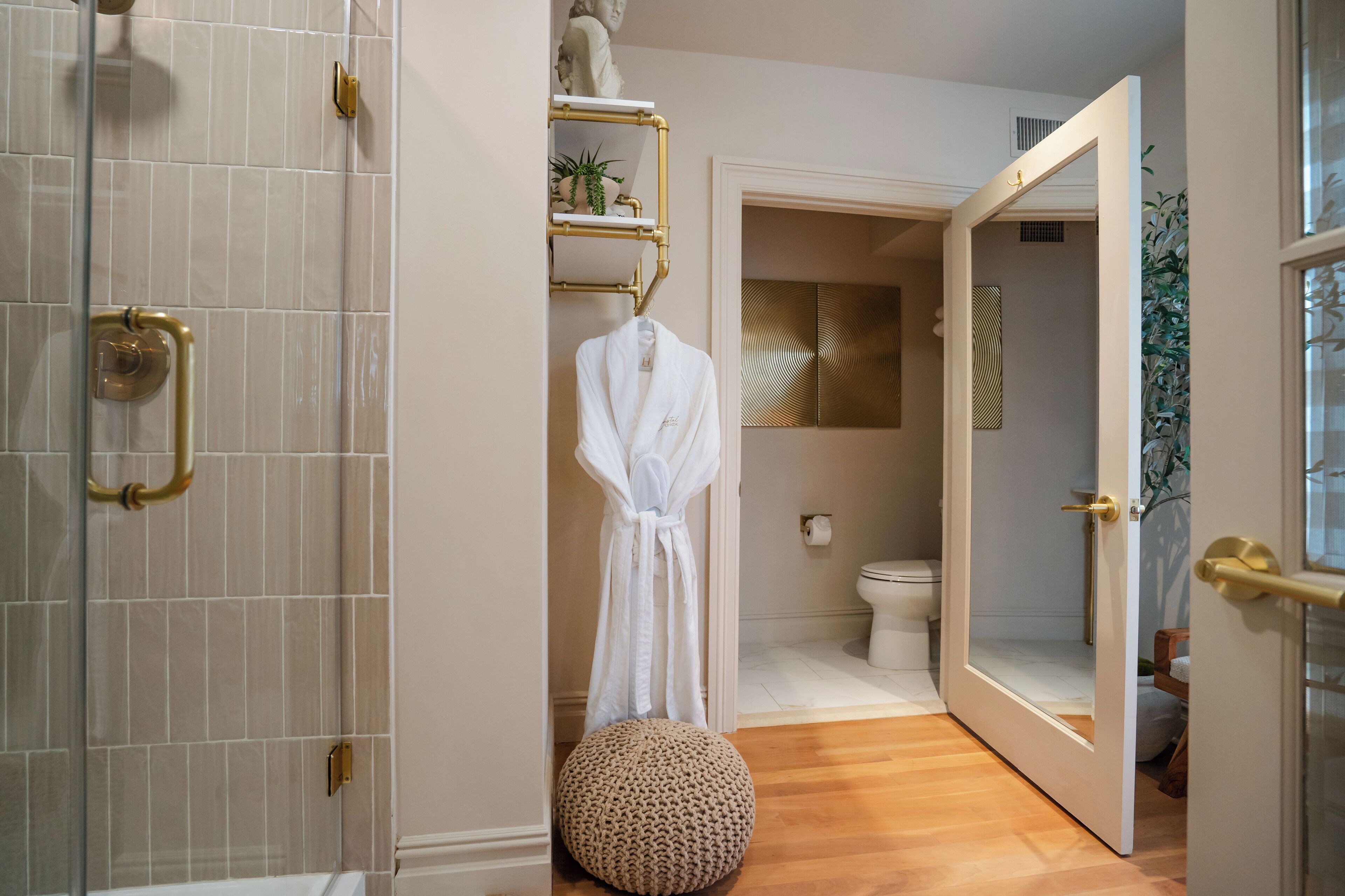 A bathroom with a shower, a toilet, and a sink. There is a robe hanging on a rack and a round ottoman on the floor.