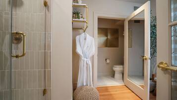 A bathroom with a shower, a toilet, and a sink. There is a robe hanging on a rack and a round ottoman on the floor.