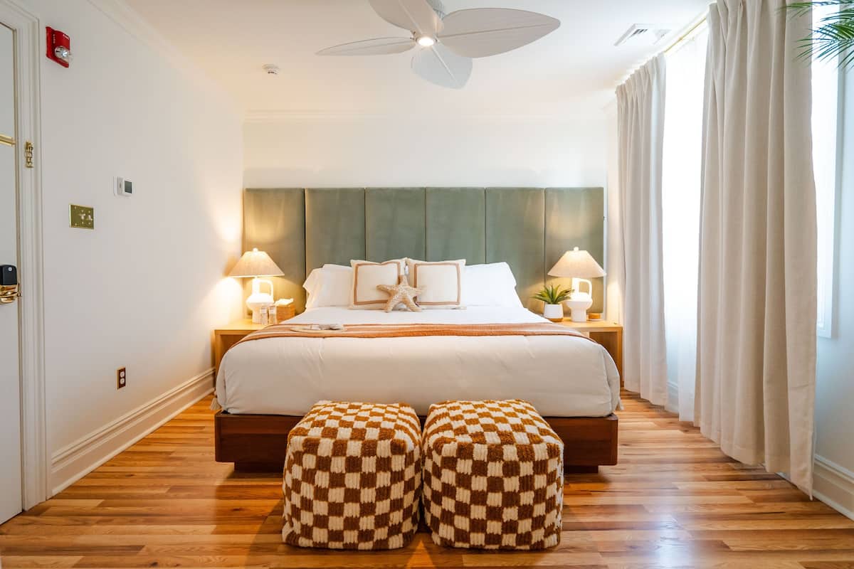 A hotel room with a large bed, two bedside lamps, a ceiling fan, and a view of the outdoors through sheer curtains.
