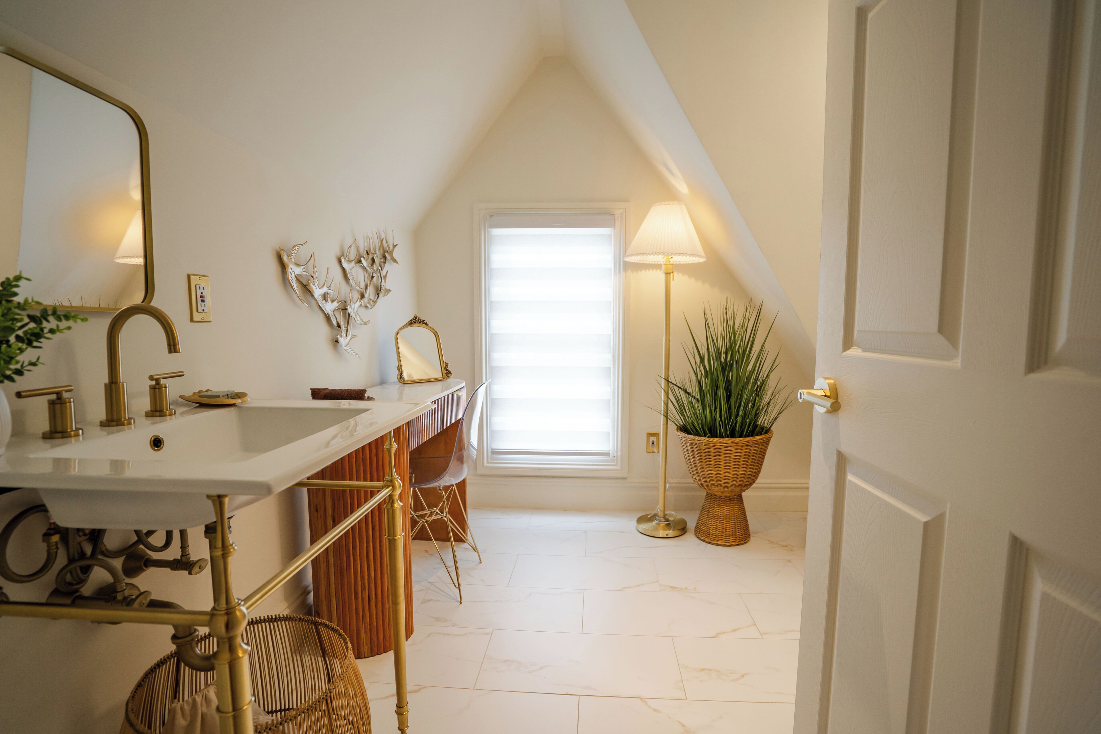 A bathroom with a white sink, gold fixtures, a mirror, a wicker chair, and a potted plant.