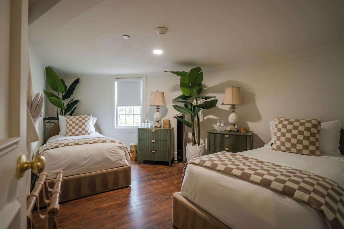 A bedroom with two beds, a window, a nightstand, a lamp, and a potted plant.