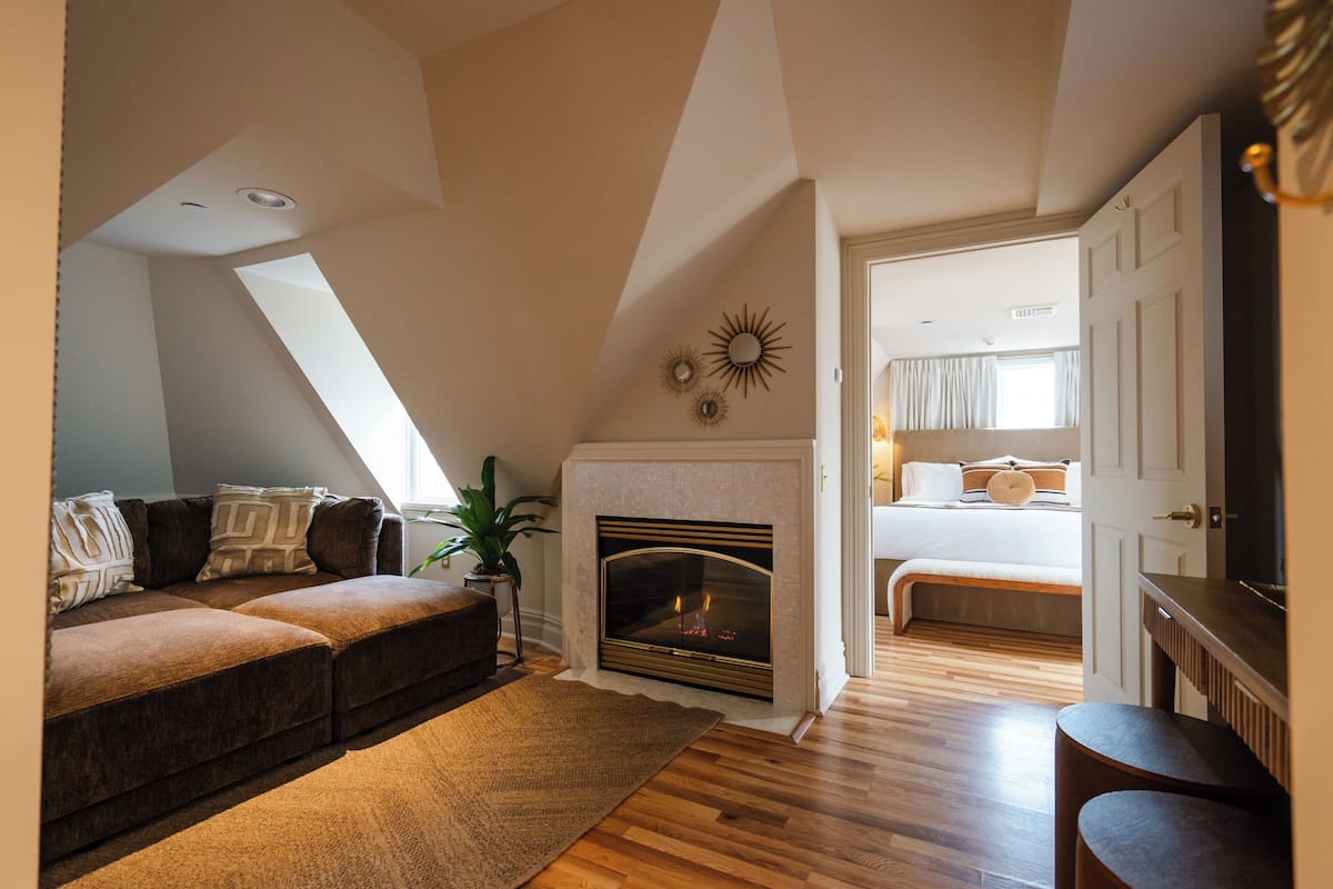 A cozy loft bedroom with a fireplace, a large window, and a view of the adjacent living room.