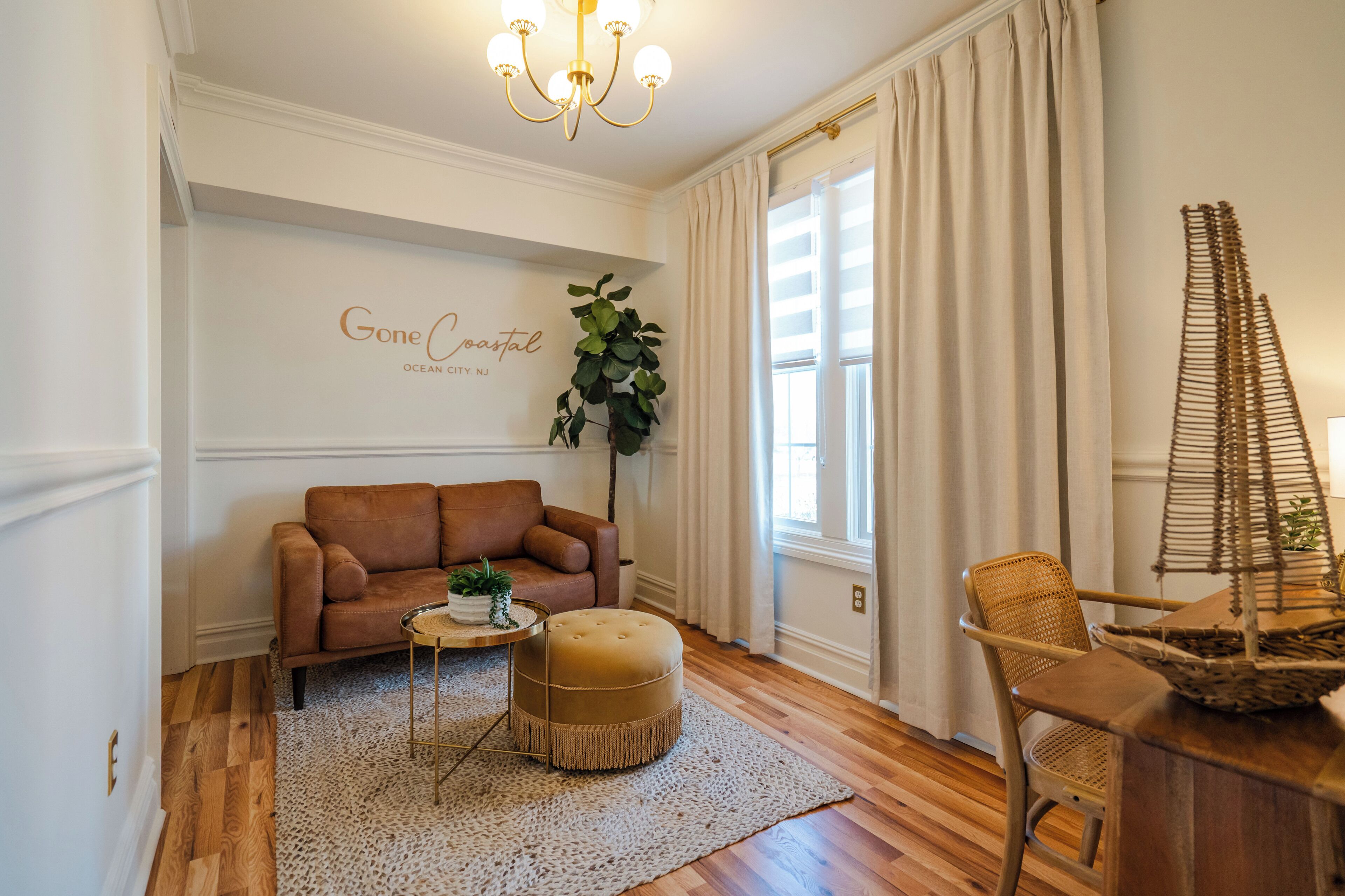 Un salon avec un canapé marron, un pouf et un fauteuil en osier. Une plante est posée sur une table d’appoint, et un mur porte l’inscription « Gone Coral ».