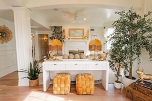 A well-lit room with a white fireplace, a large potted plant, and a white table with two woven stools.