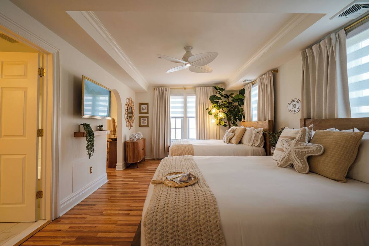 A hotel room with two beds, a ceiling fan, a TV, and a large window with curtains.