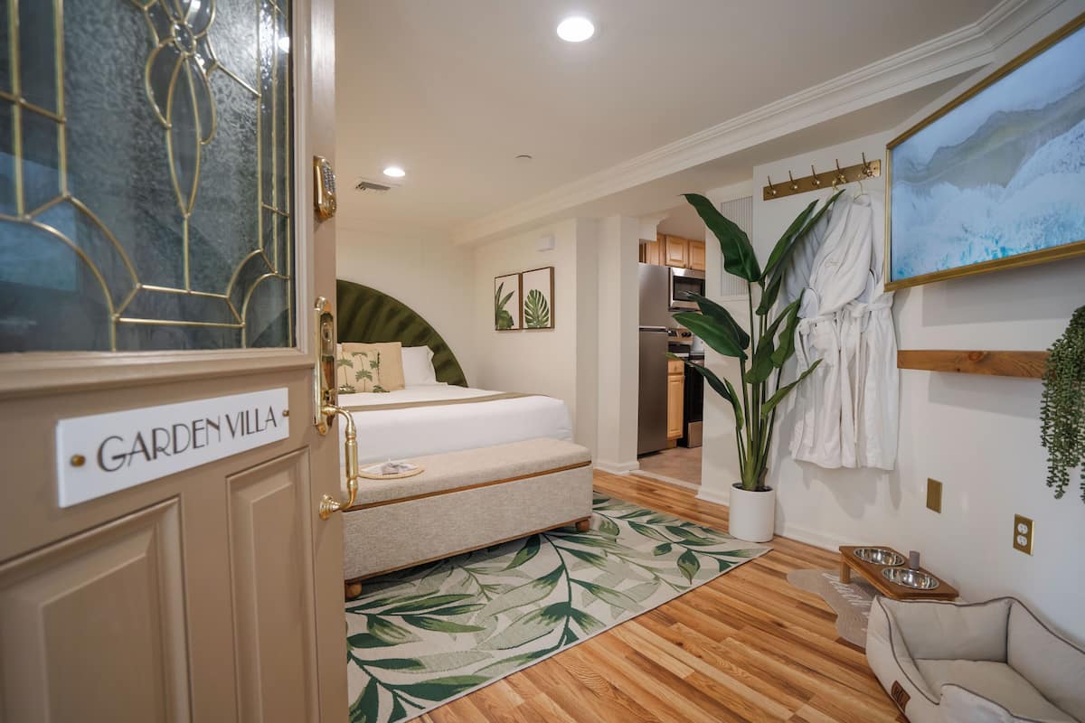 A bedroom with a bed, a bedside table, a large potted plant, a painting, and a wooden floor.