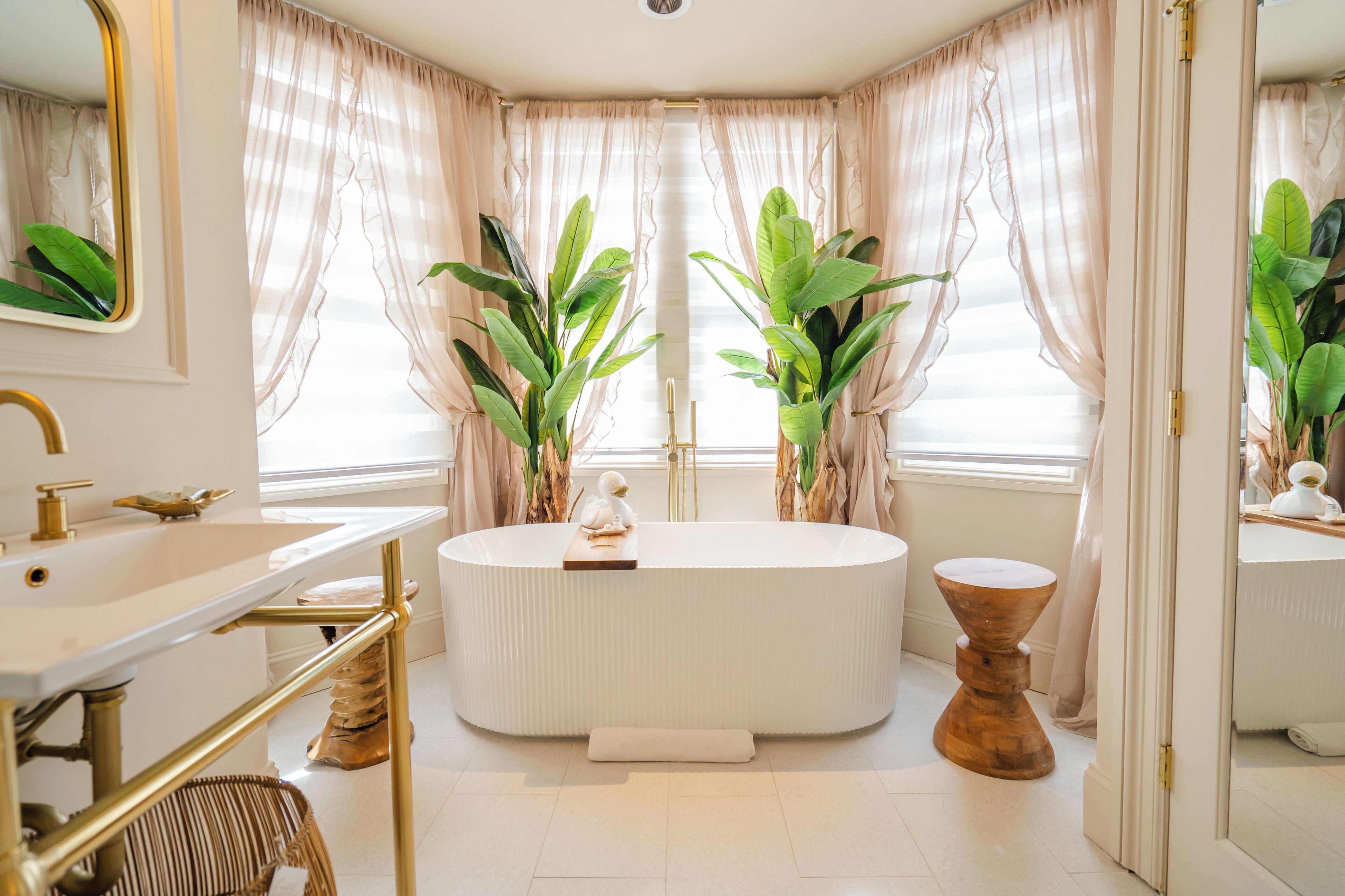 A modern bathroom with a freestanding bathtub, a vanity with a gold faucet, and large windows with sheer curtains.