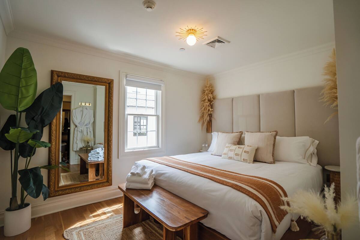 A bedroom with a large bed, a wooden bench, a mirror, and a potted plant.