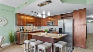 A modern kitchen with wooden cabinets, a stainless steel refrigerator, a microwave, and a large island with bar stools. - Nautical Nest at Laketown Wharf 1019 | Beach Access, Gym, Parking Onsite, 5 Pools (Panama City Beach)