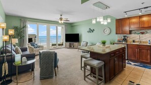 A spacious living room with a large TV, comfortable seating, a kitchen with wooden cabinets, and a balcony with a view. - Nautical Nest at Laketown Wharf 1019 | Beach Access, Gym, Parking Onsite, 5 Pools (Panama City Beach)