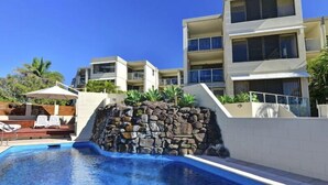 Une piscine agrémentée d’une cascade en pierre, entourée d’un mur blanc et d’un immeuble à plusieurs étages doté de balcons.