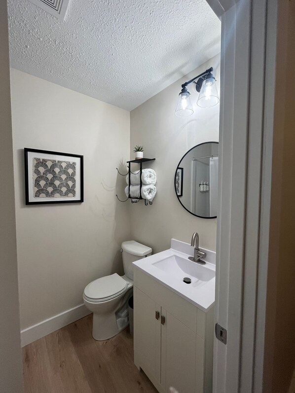 A small, clean bathroom with a toilet, sink, and mirror. There is a framed picture on the wall and a small shelf with towels.
