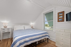 Quarto com cama, mesas de cabeceira, cômoda, janela com persianas e televisão fixada na parede.