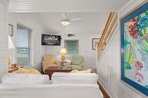 A hotel room with a bed, two chairs, a table with a lamp, a ceiling fan, a TV displaying 'Coastal Carolina Vacations and Sales', and a large painting on the wall.