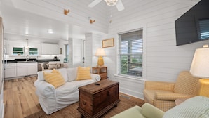 A spacious living room with a white sofa, a wooden chest of drawers, and a checkered armchair. A television is mounted on the wall, and a window with blinds is visible. - Nags Head 364: Pet Friendly, Newly Remodeled, Oceanside, Short Stays Anytime! (Nags Head)