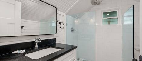 Un baño moderno con un espejo grande, una encimera negra, un lavabo blanco y una zona de ducha con mampara de cristal.