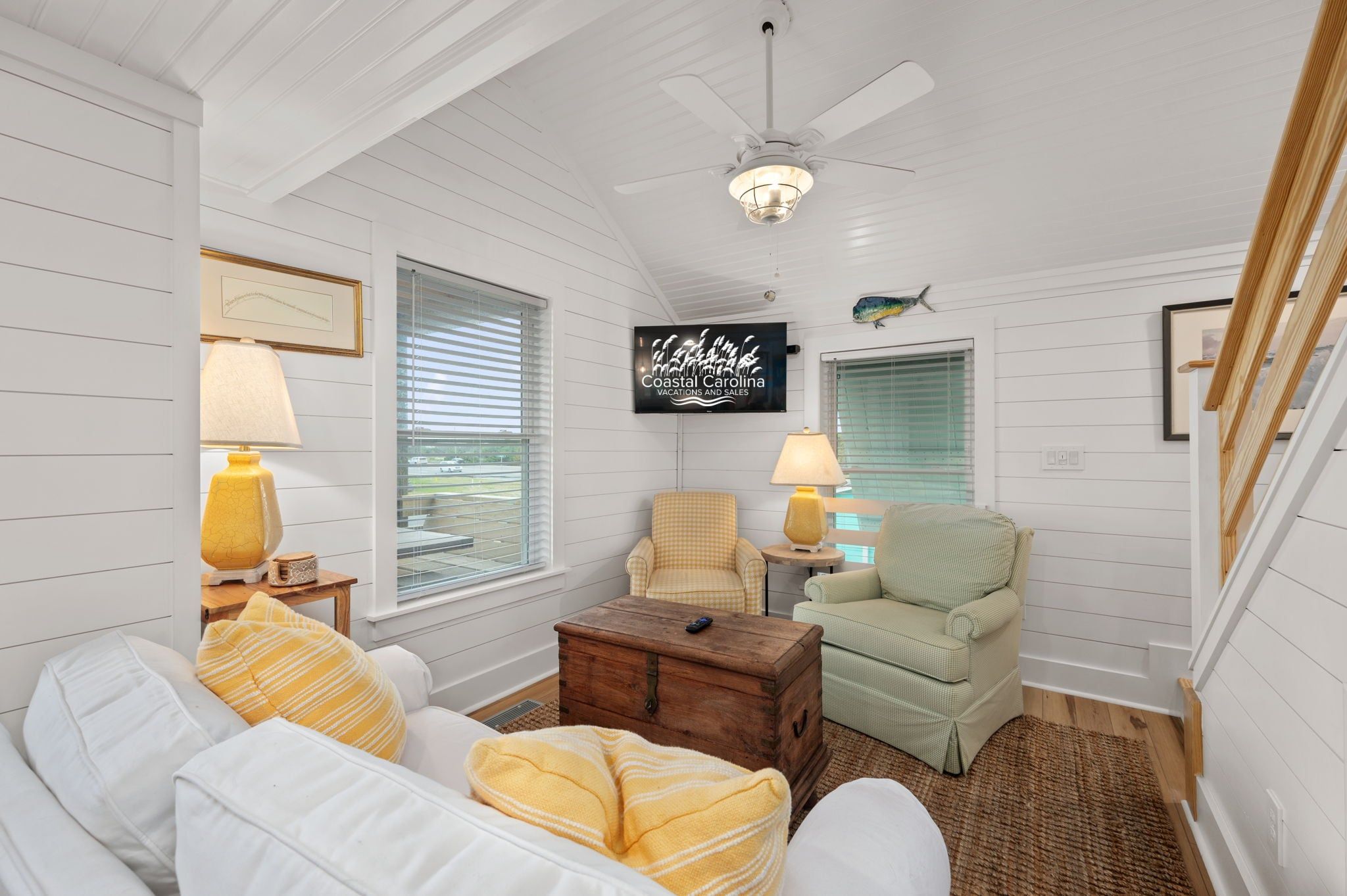 Une pièce accueillante aux murs recouverts de lambris blancs, dotée d’un ventilateur de plafond, d’un téléviseur diffusant des publicités pour « Coastal Carolina Vacations and Sales » et d’une fenêtre avec des stores.