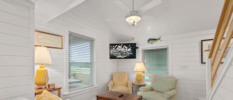 A cozy room with white paneled walls, a ceiling fan, a television displaying 'Coastal Carolina Vacations and Sales', and a window with blinds.