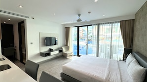 A modern hotel room with a large bed, a TV mounted on the wall, a desk, and a view of a pool through the windows. - Private studio at Twin Sands Resort (Patong)
