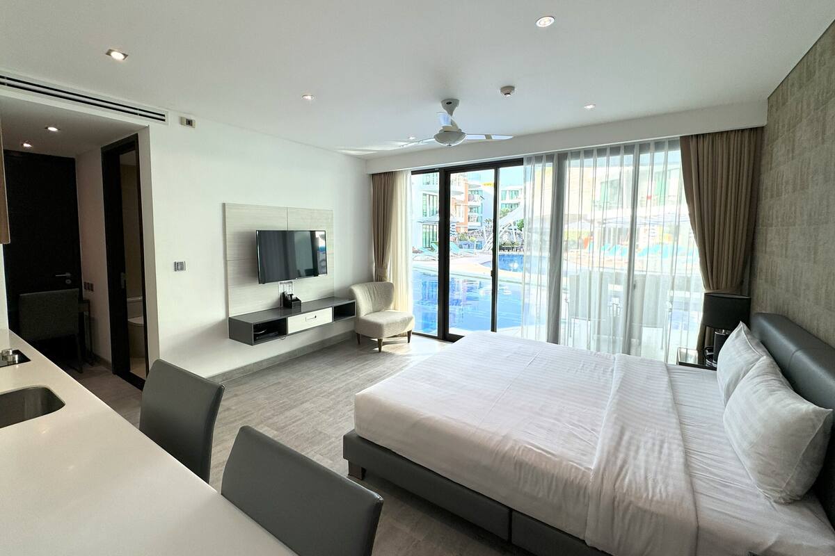 Quarto de hotel moderno com uma cama grande, uma TV na parede, uma escrivaninha e vista para uma piscina pelas janelas.