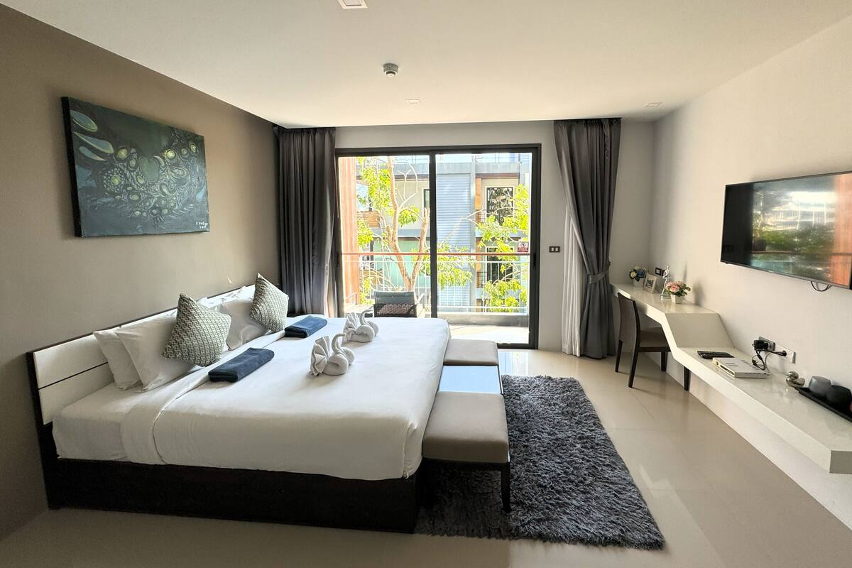 Ett modernt hotellrum med en stor säng, ett skrivbord, en TV och en balkong med utsikt över grönområden.