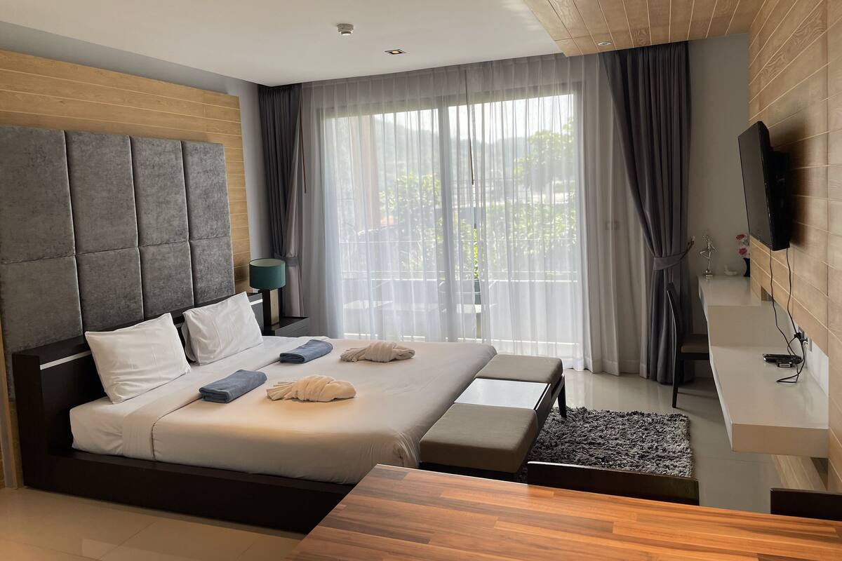 Ett modernt hotellrum med en stor säng, ett träbeklätt sänggavel, en platt-TV och utsikt över grönområden utanför.