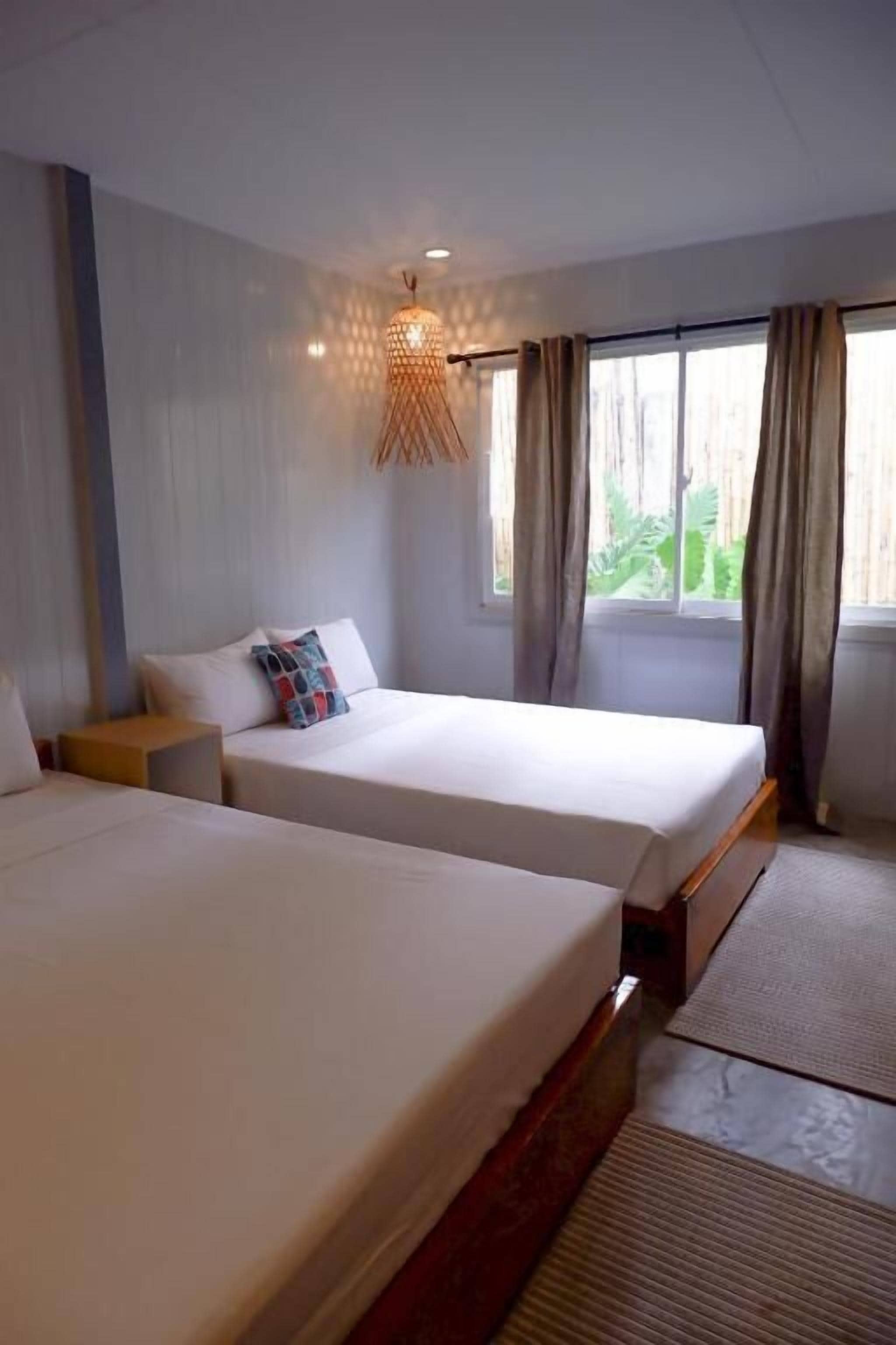 A hotel room with two beds, a bedside table, a hanging light fixture, and a window with curtains.