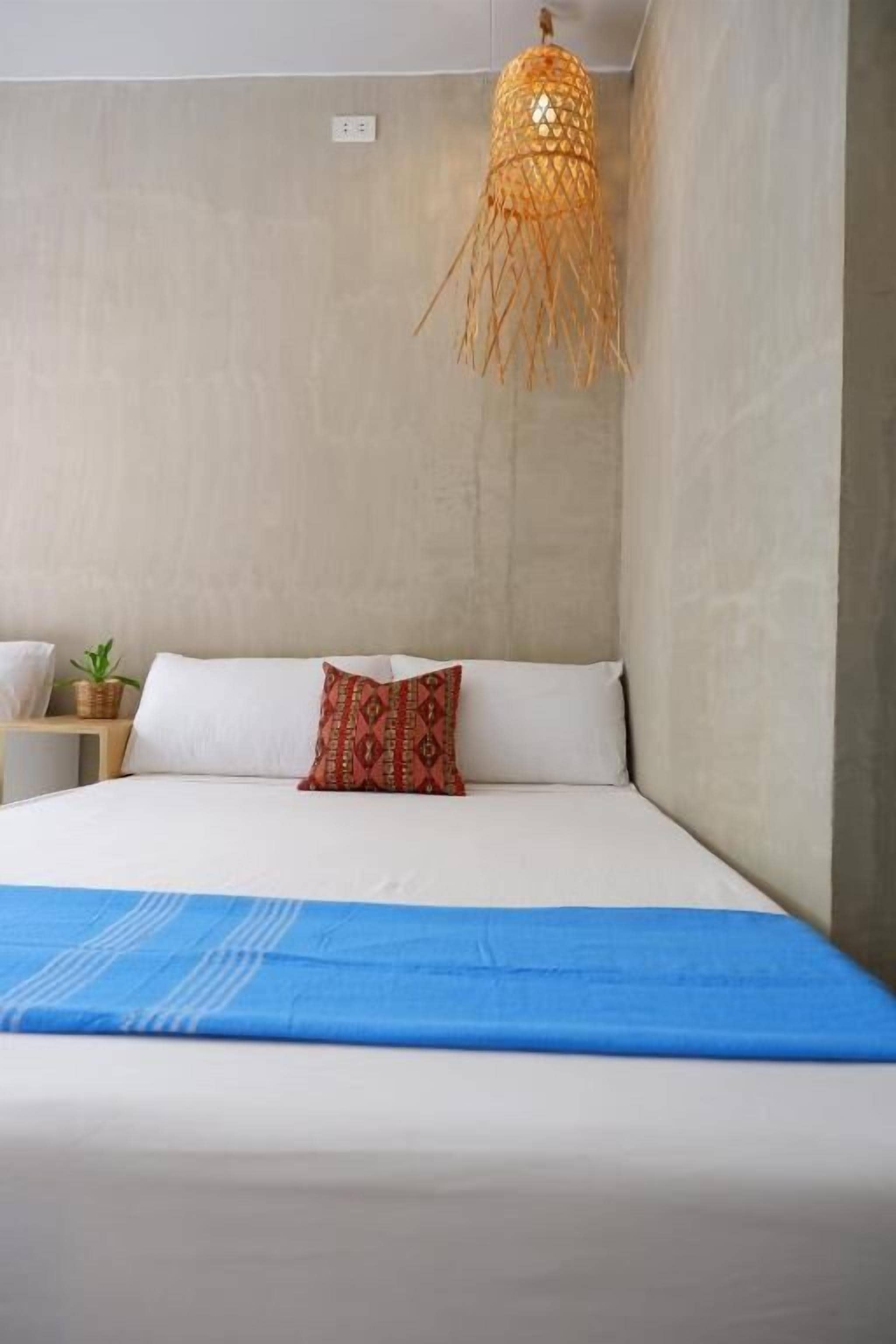 A bedroom with a bed, a red patterned pillow, a blue bedspread, a woven hanging light fixture, and a small bedside table with a plant.