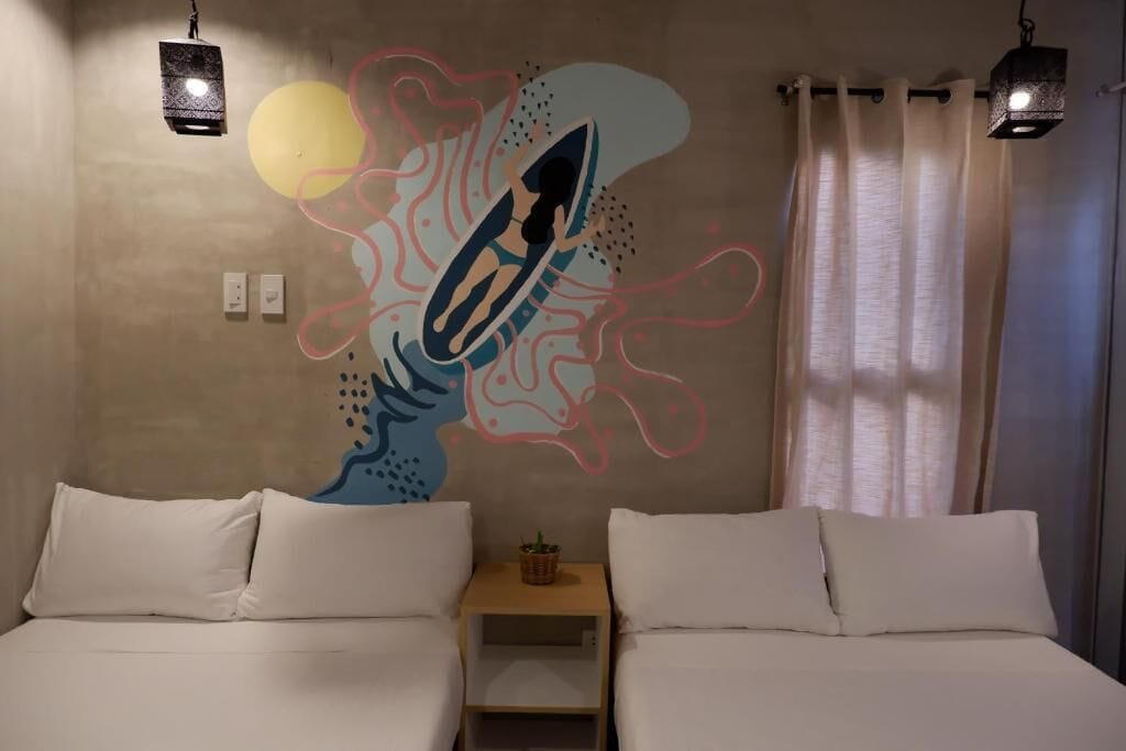 A room with two beds, a wall mural, a small table with a plant, and two hanging lamps.
