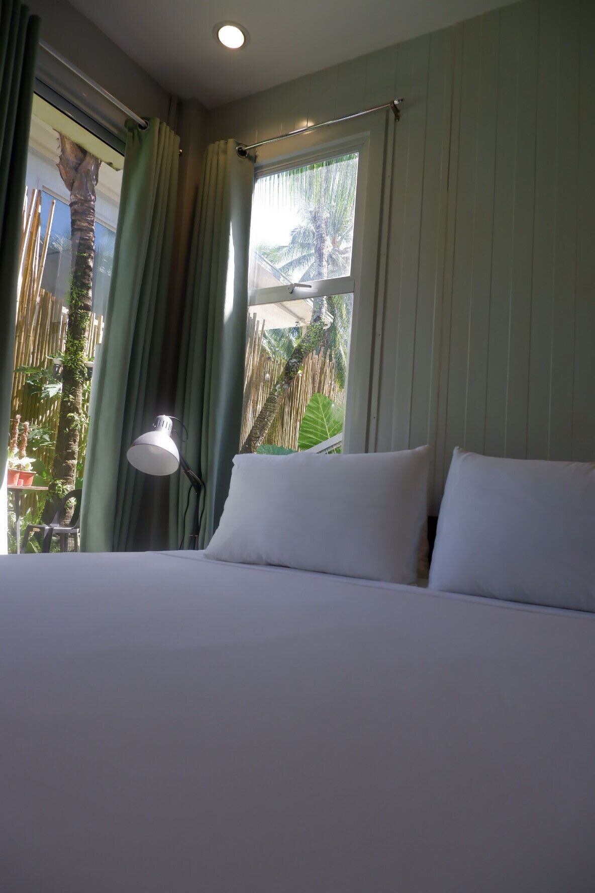 A hotel room with a bed, a window with curtains, and a view of a palm tree and fence.